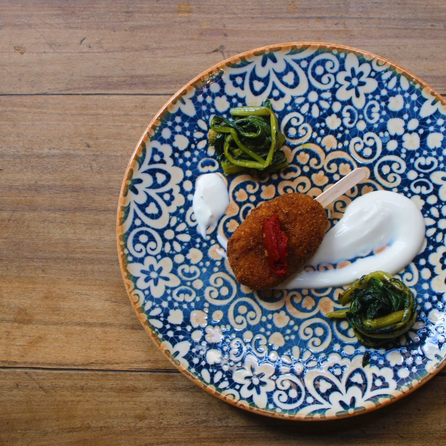 Cibo fritto su uno stecco, servito con verdure e salsa bianca su un piatto decorato con motivi blu e bianchi appoggiato su un piatto di legno.
