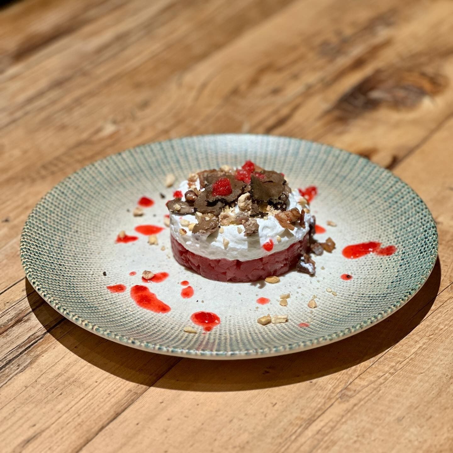 Dessert su un piatto: strati di ingredienti rossi, bianchi e marroni con glassa rossa e noci su un tavolo di legno.