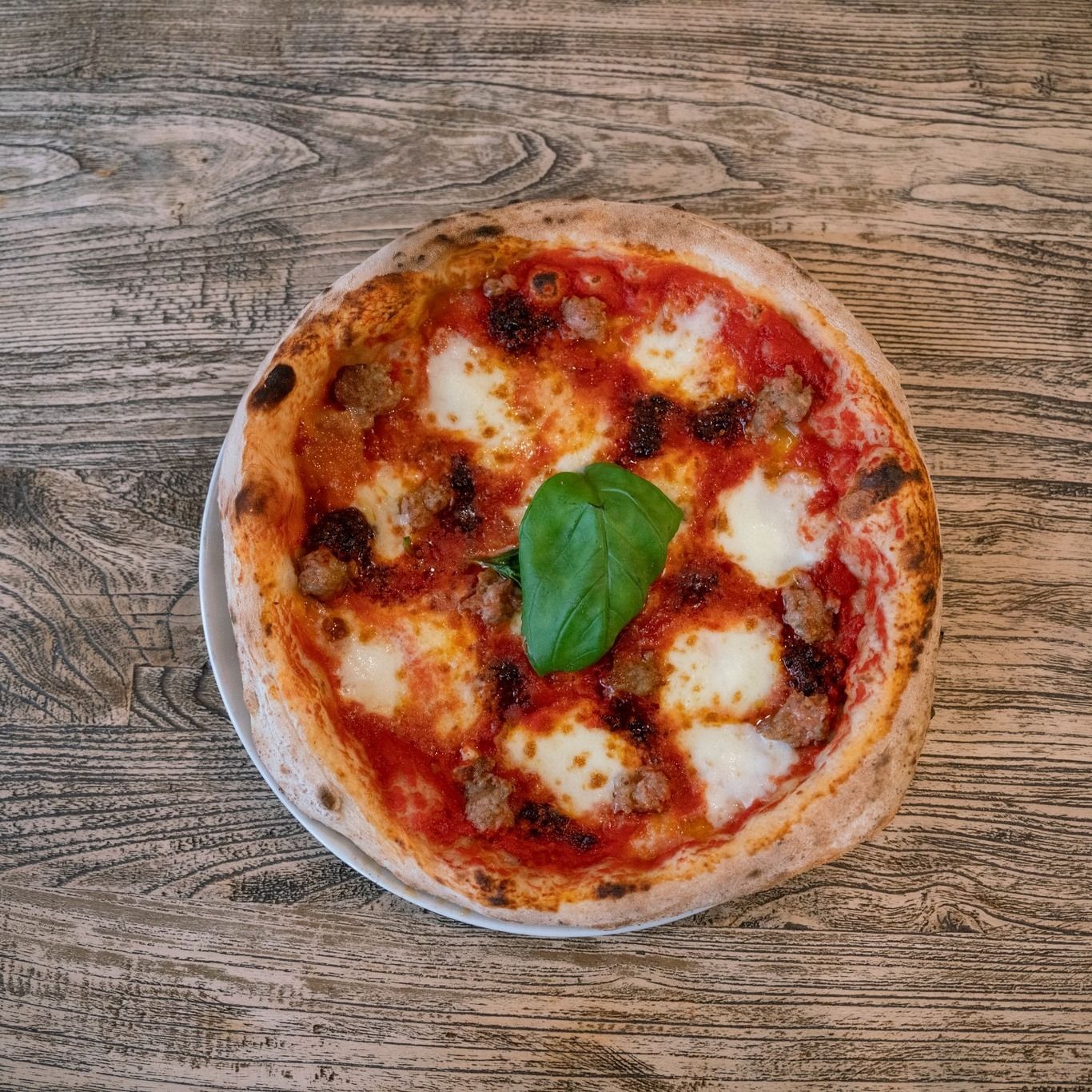 Pizza su un piatto bianco, appoggiata su una superficie di legno, con formaggio, salsiccia e una foglia di basilico.