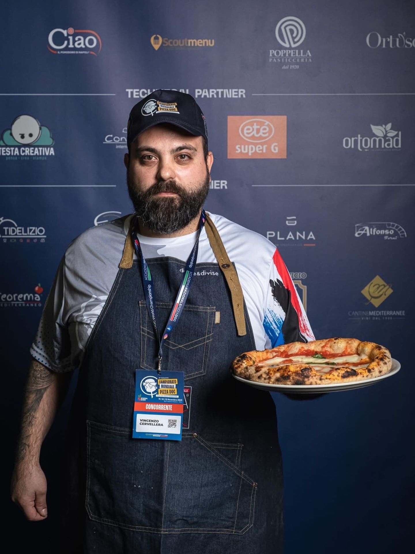 Uomo con in mano una pizza, con grembiule e berretto da baseball, in piedi davanti a uno sfondo blu con loghi.