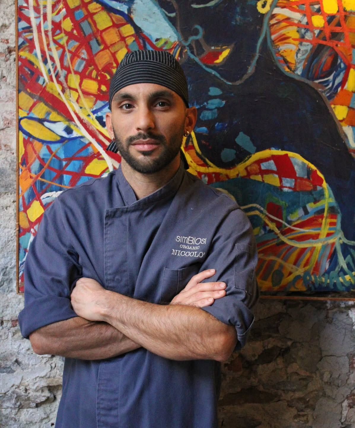 Uno chef in uniforme blu, con le braccia incrociate, è in piedi davanti a un dipinto astratto colorato.