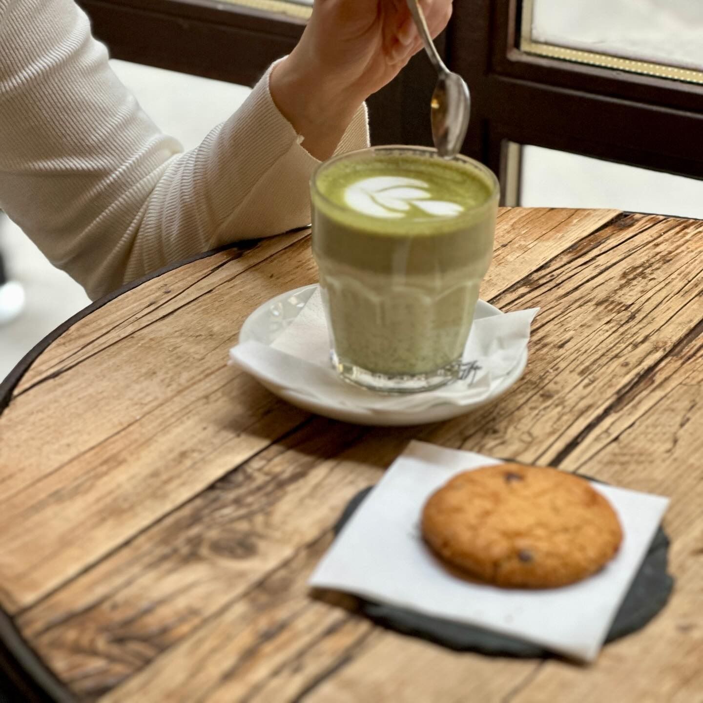 Un matcha latte con latte art e un biscotto su un tavolo di legno. Una persona mescola la bevanda con un cucchiaio.