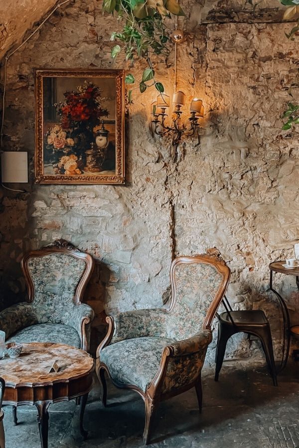 Due poltrone decorate e un tavolino in una stanza con una parete in pietra e un lampadario. Sopra, un dipinto di fiori.