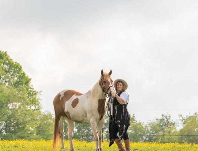 A woman is standing next to a brown and white horse in a field of yellow flowers.