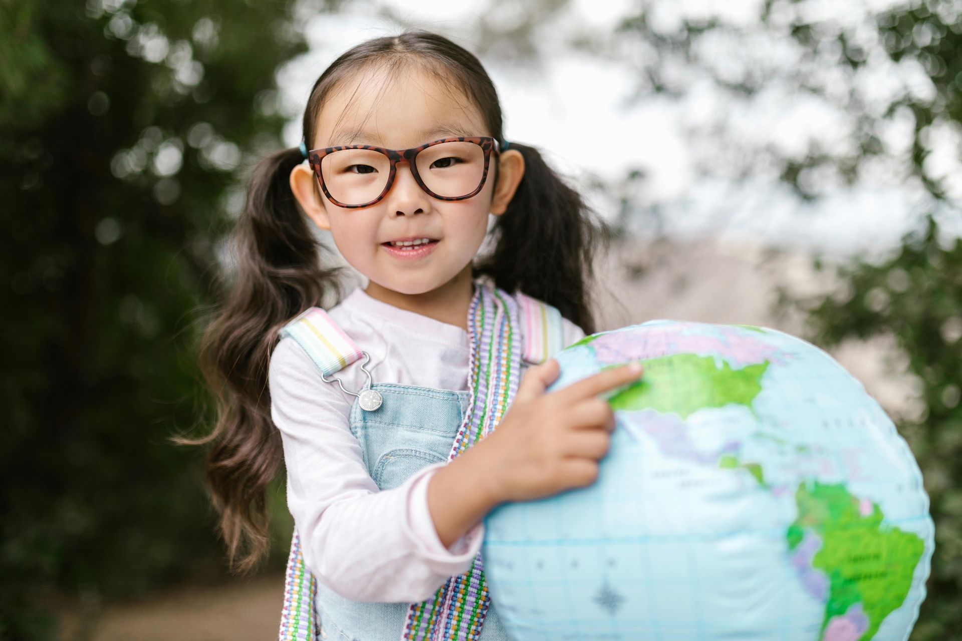 A little girl wearing glasses is holding a globe and pointing at it.