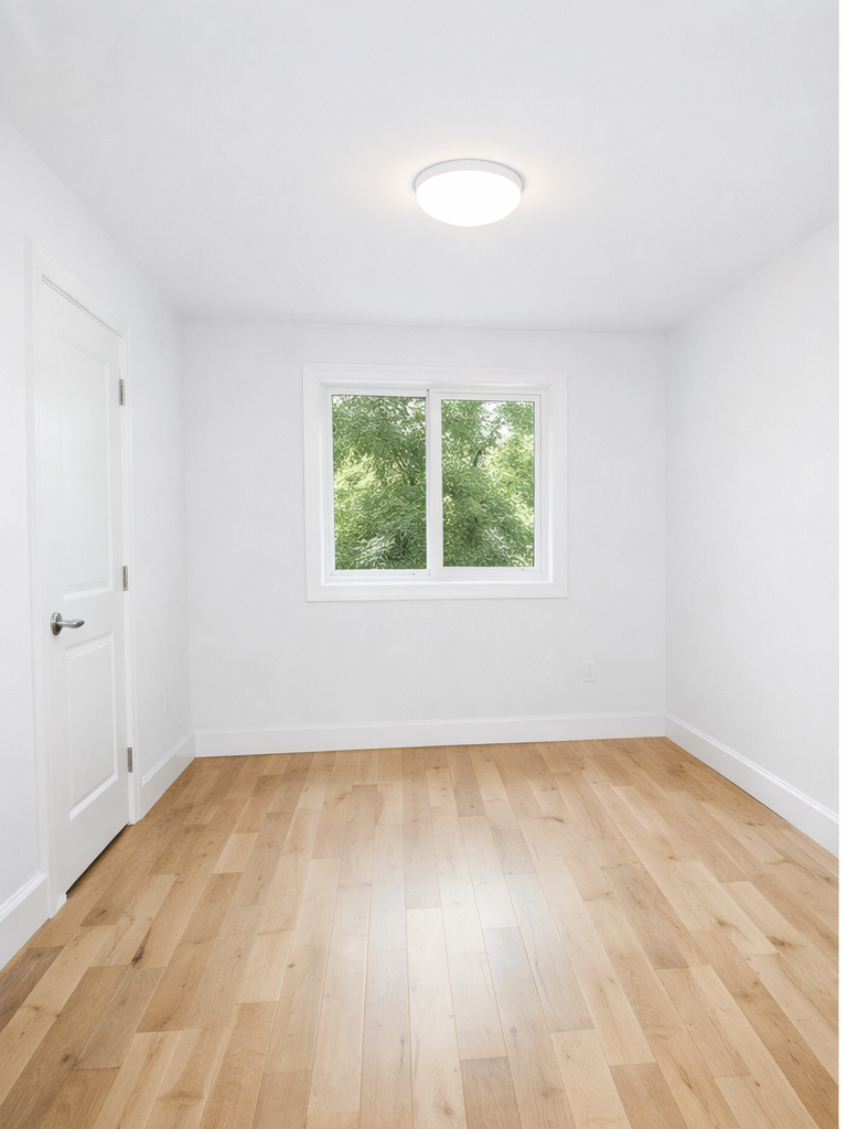 An empty, minimalist room with white walls, light wood flooring, a single window facing trees, and a ceiling light.
