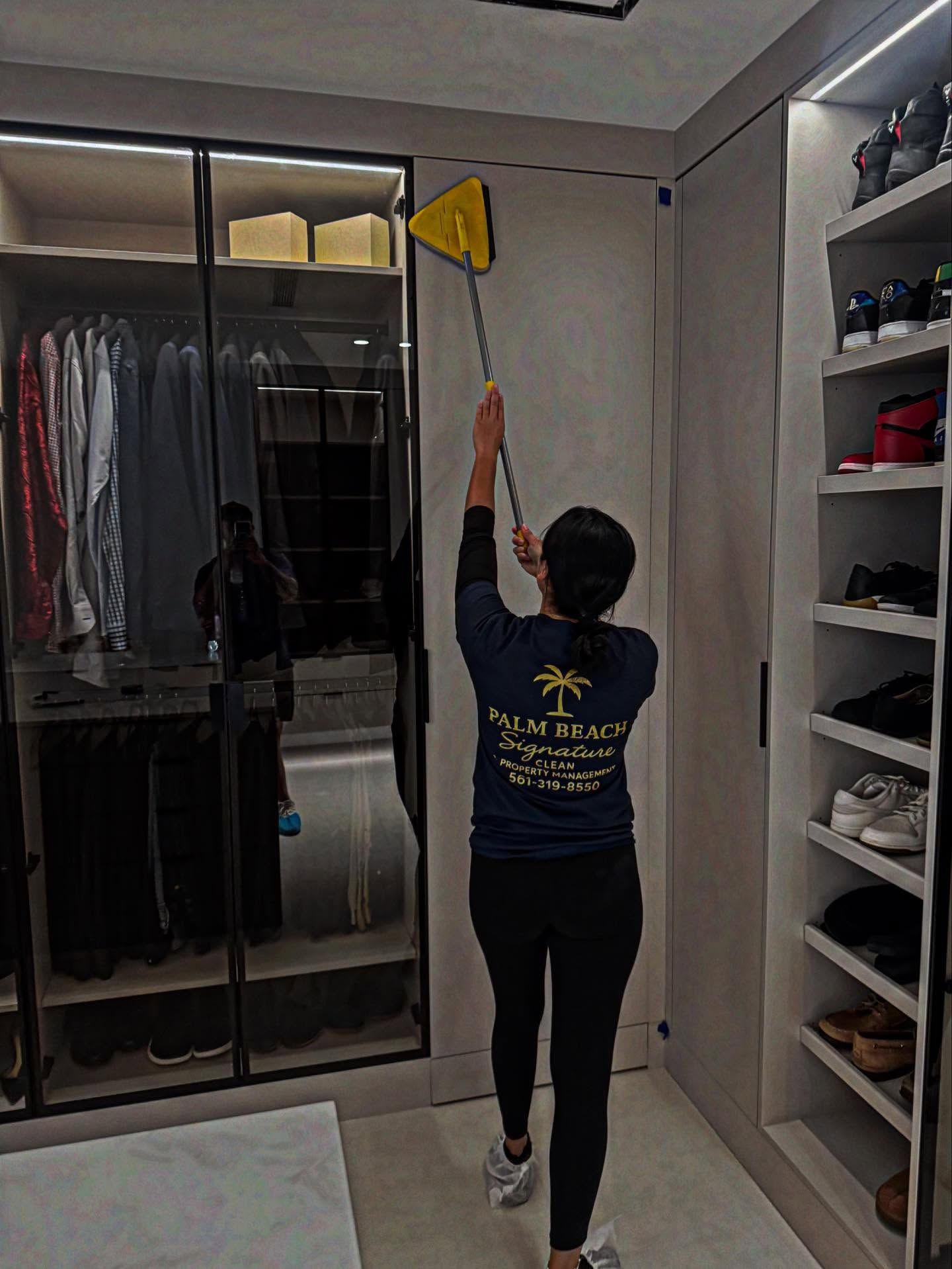 Woman in a closet cleaning high wall with a duster. She is wearing a shirt with a palm tree logo.