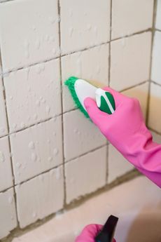 A person wearing pink gloves scrubs bathroom tiles with a green and white brush.