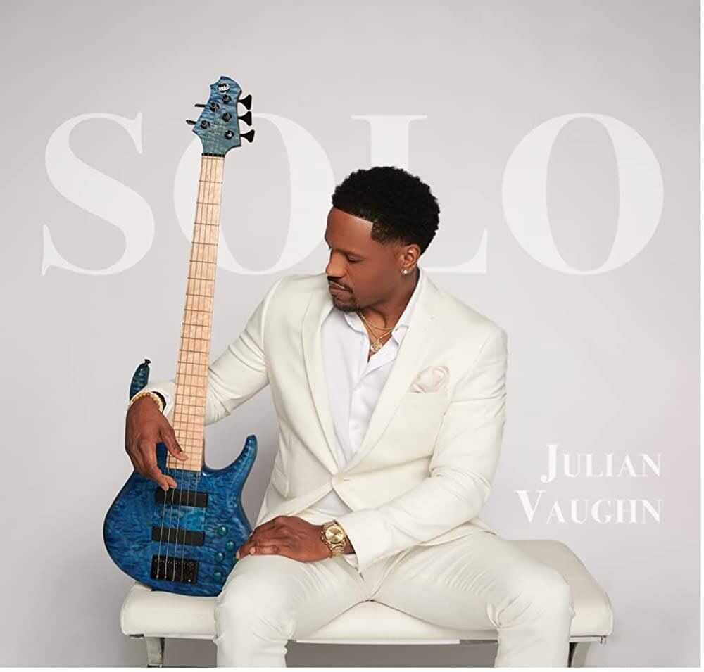 Julian Vaughn, in white suit, holding blue bass guitar, sitting on white bench, gazing right.