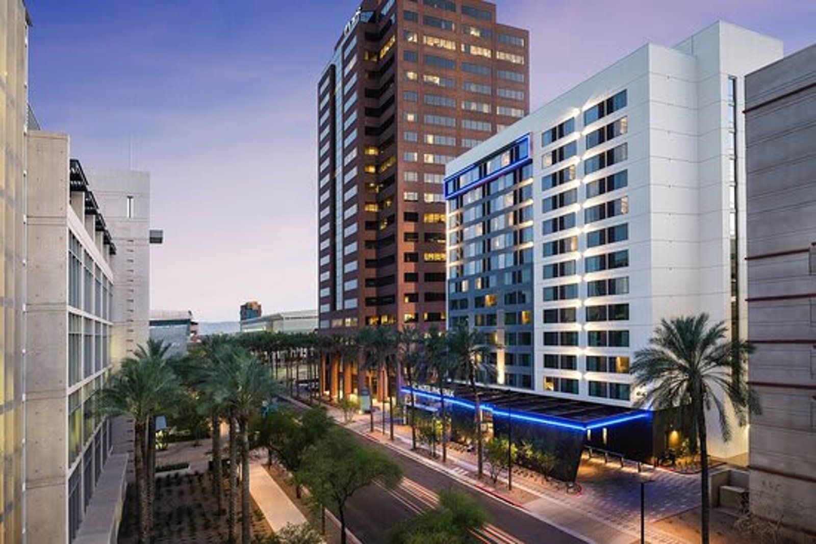 Modern hotel exterior in a city setting, with palm trees, blue accents, and a tall brown building behind it.