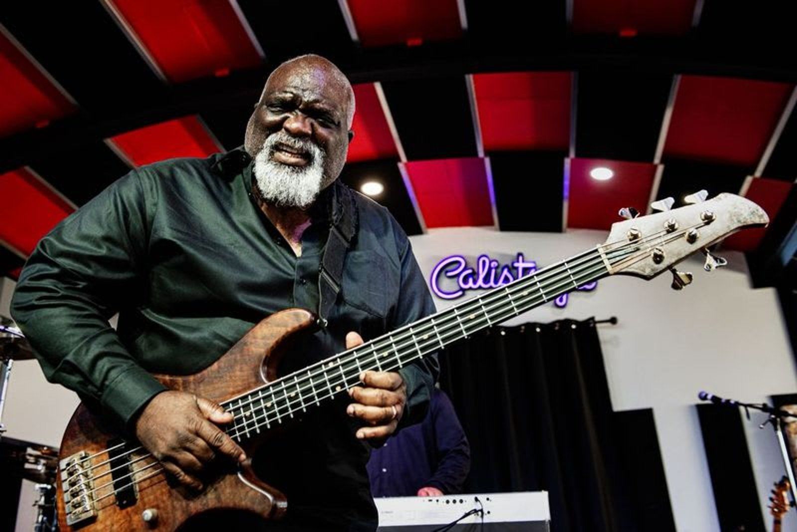 Man with a white beard playing a brown bass guitar at a music studio, black shirt, Calisto sign.