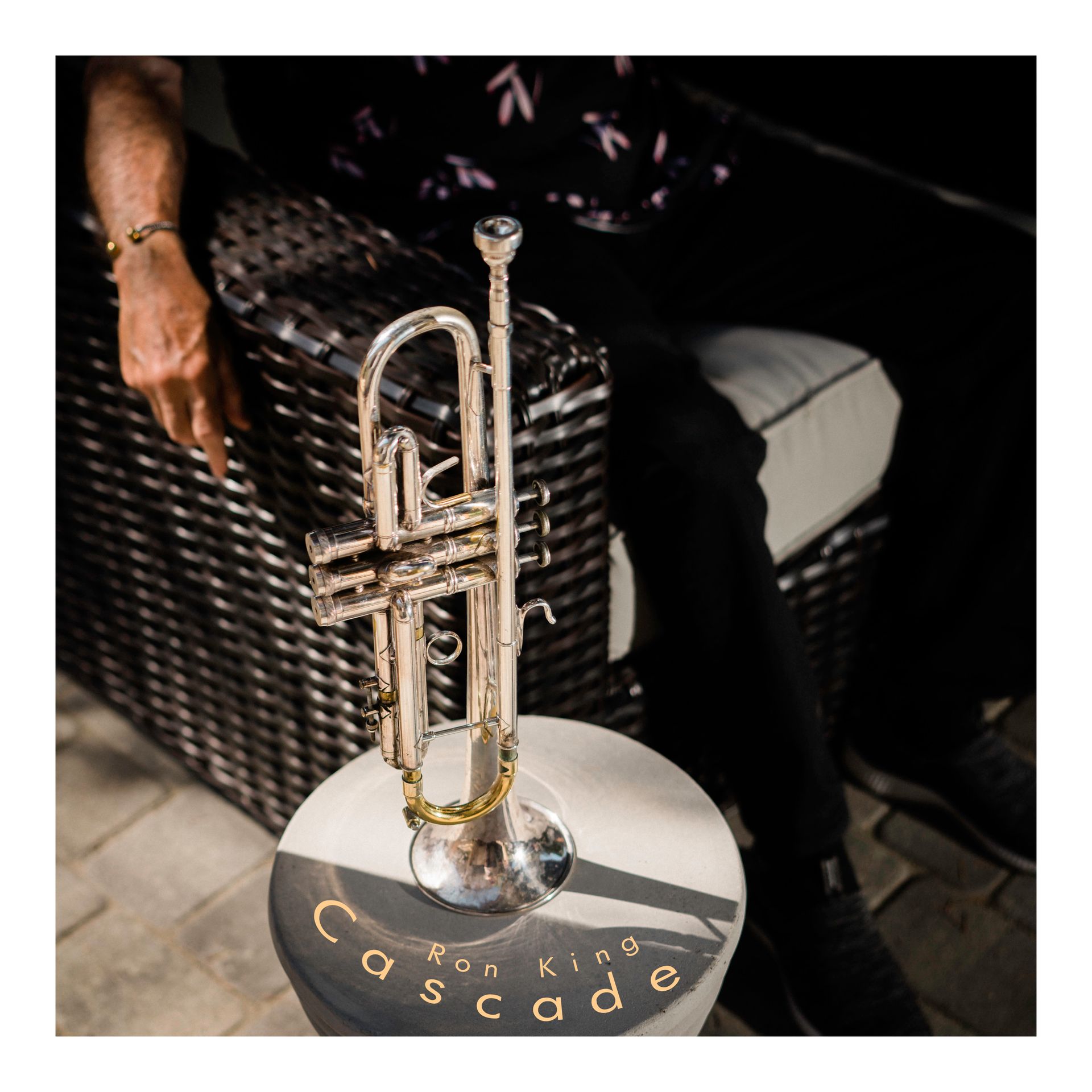 A silver trumpet on a round table, near a seated person wearing black pants and a patterned shirt.