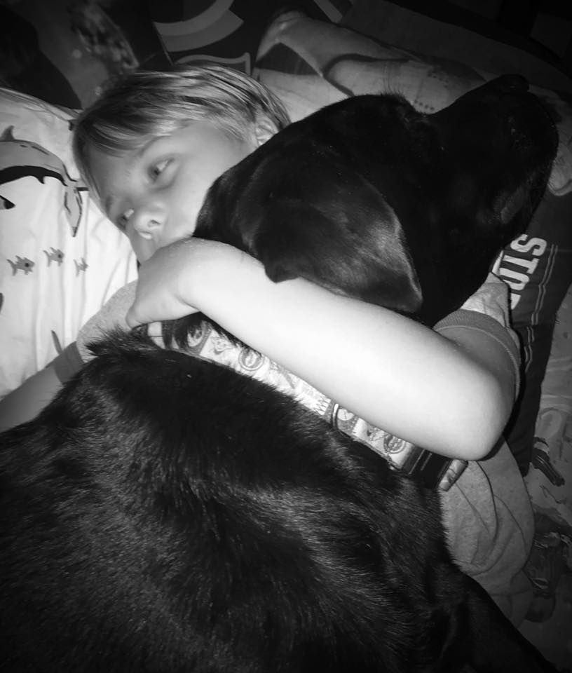 A black and white photo of a child hugging a black dog
