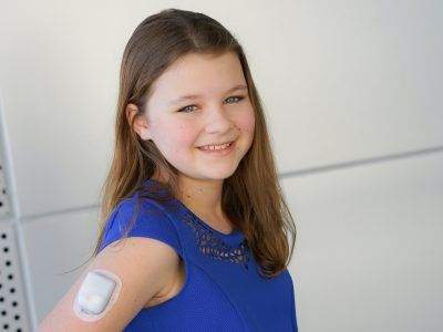 A young girl in a blue dress with a CGM on her arm.
