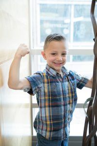 A young boy in a plaid shirt is flexing his muscles in front of a window.