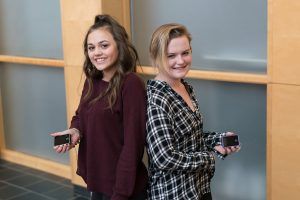 Two young women are standing back to back holding cell phones.