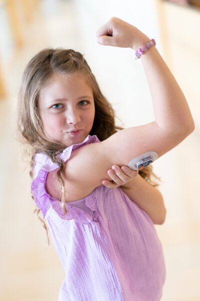 Young girl smiling and flexing her arm that has a CGM device on it.