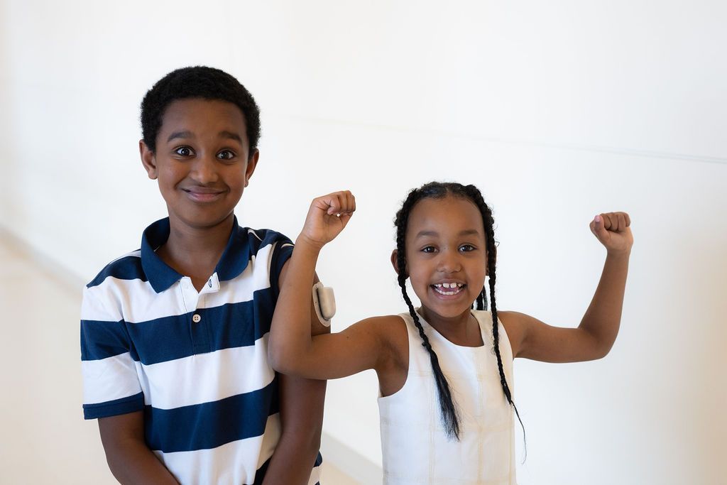 Boy with CGM on arm standing with little sister who is posing with arms flexed.