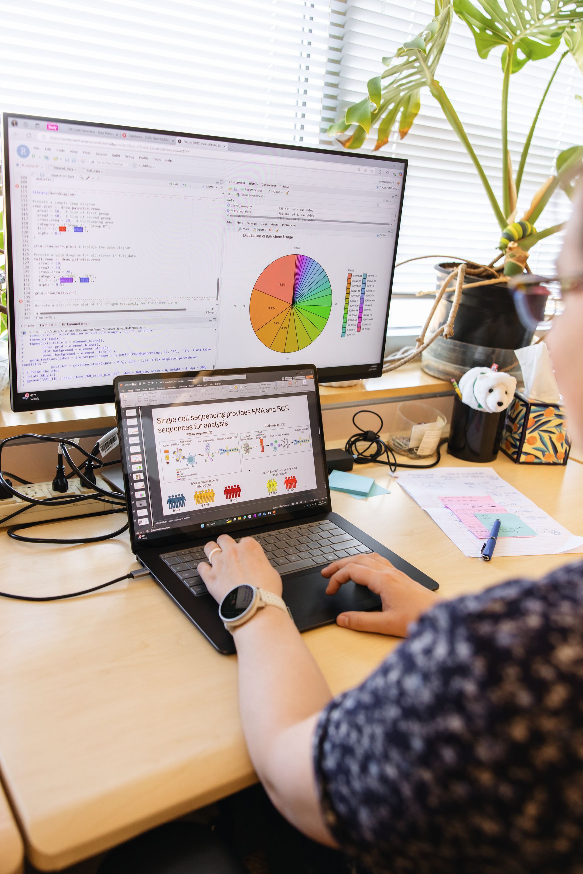 Researcher's hands typing on laptop with large screen of data in the background.