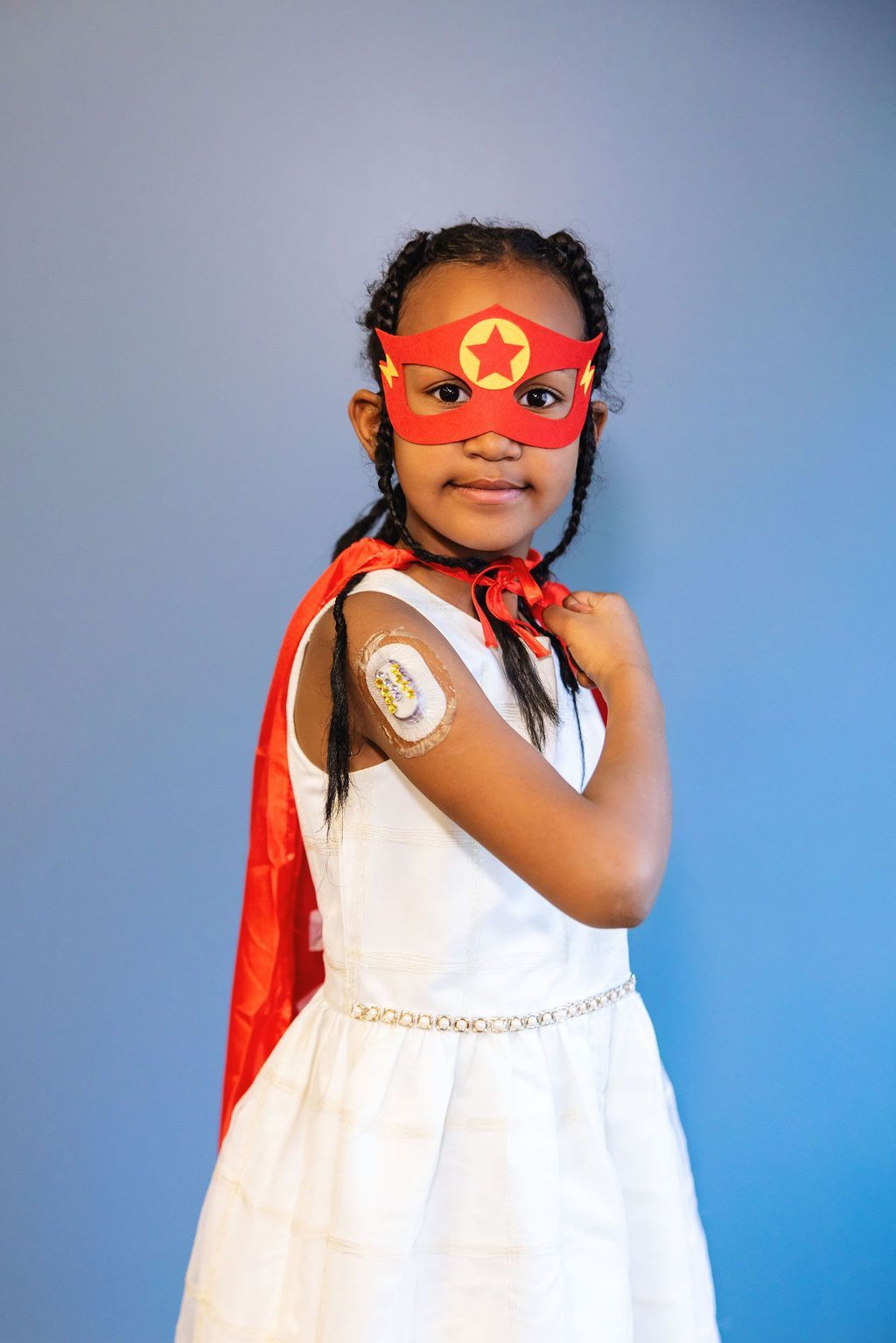Young female T1D patient in a superhero mask and cape, showing her CGM device on her arm