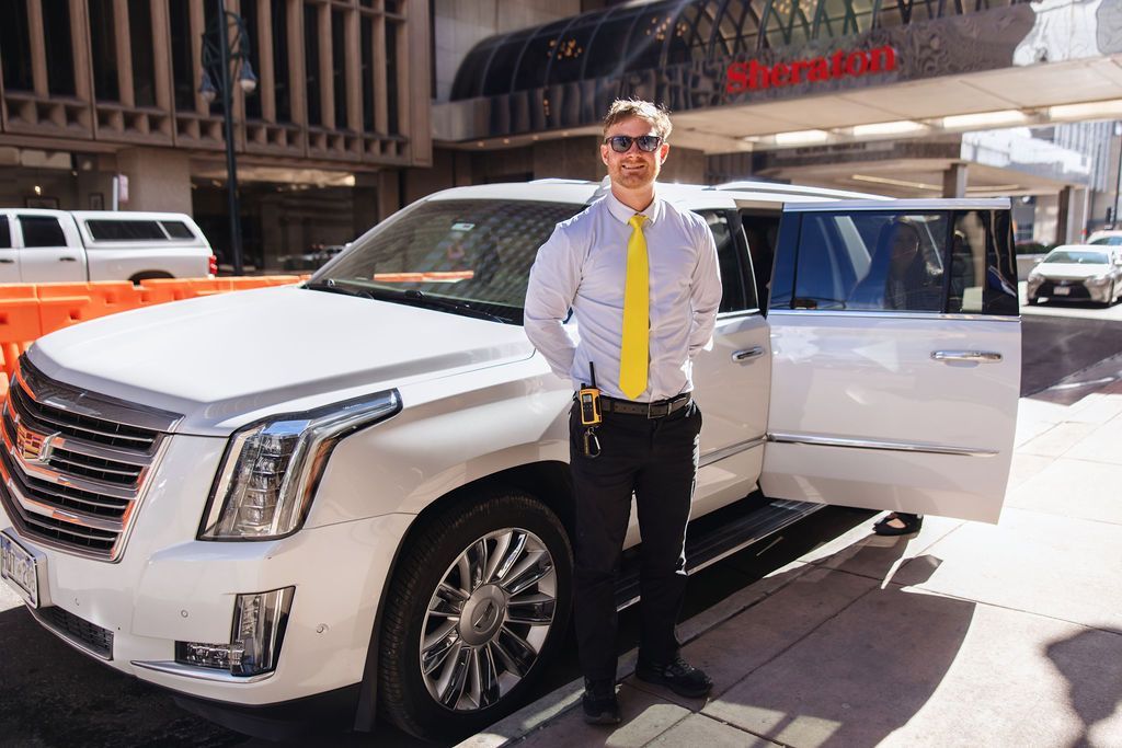 Hotel valet in tie waiting to help passengers into a white Cadillac Escalade.