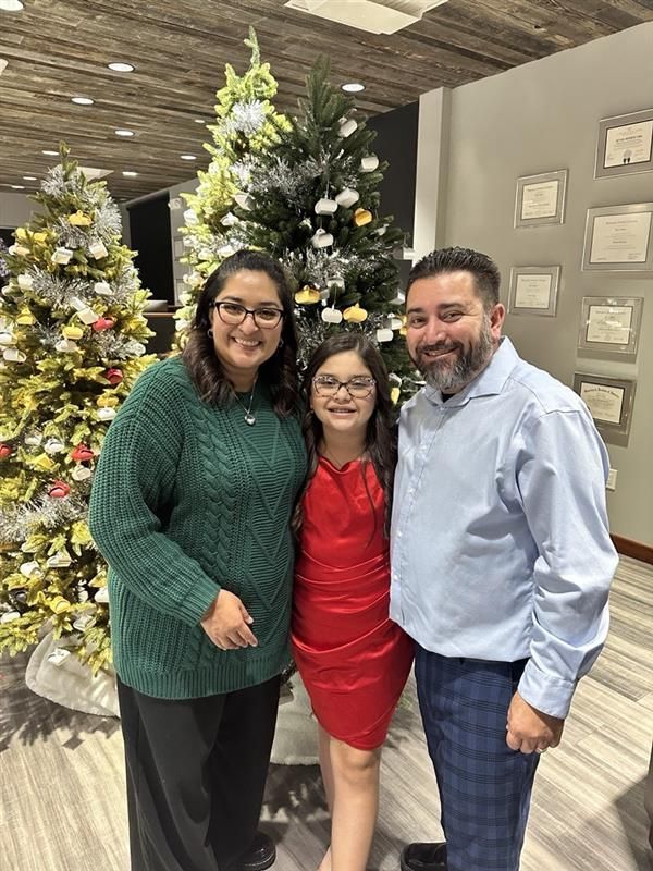 Three people smiling in front of decorated Christmas trees indoors.