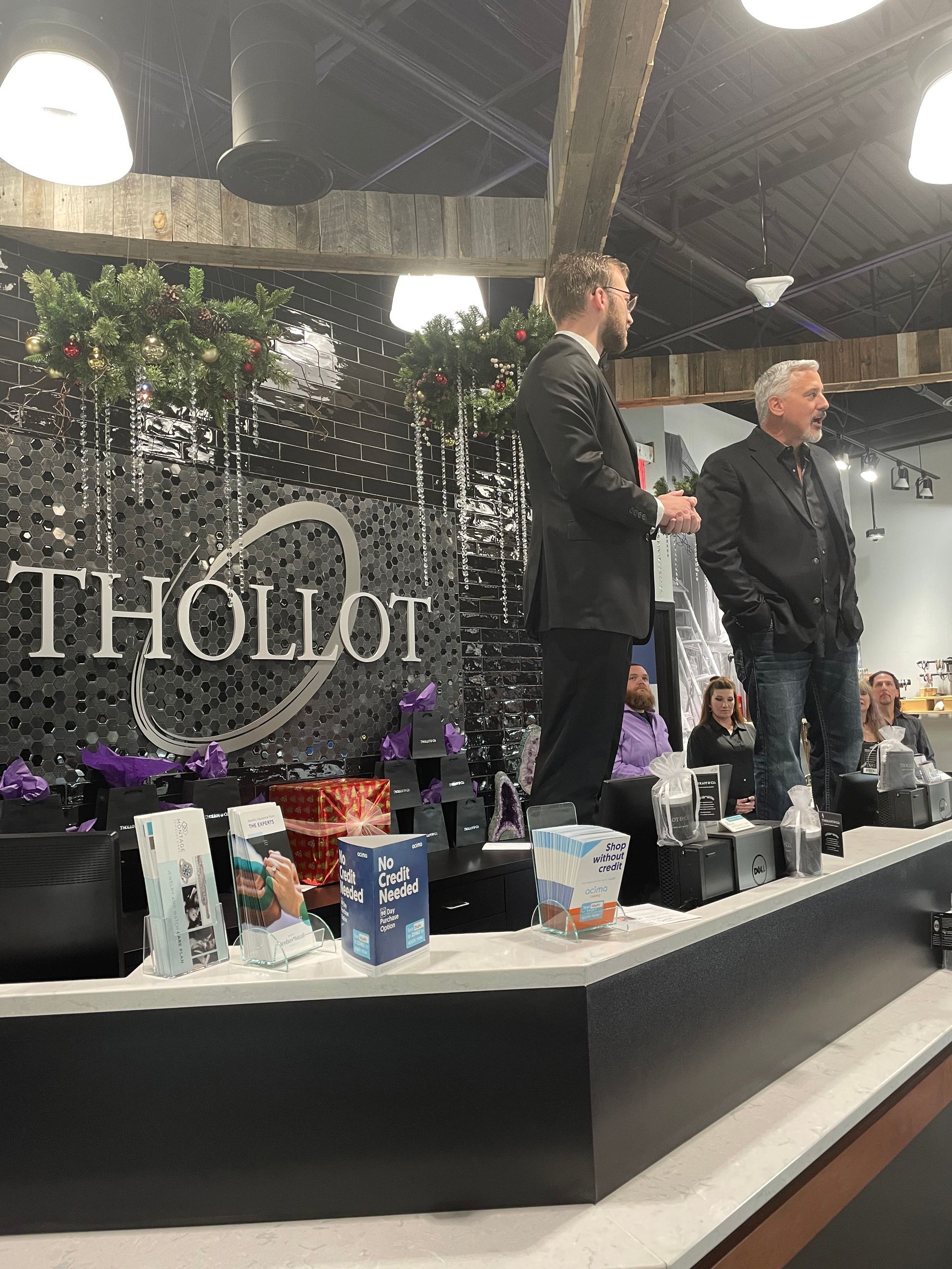 Two men standing and speaking on a stage decorated with a shiny black backdrop with the THOLLOT logo.