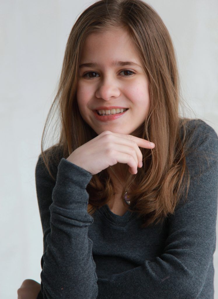 A young girl wearing a grey shirt is smiling with her hand on her chin
