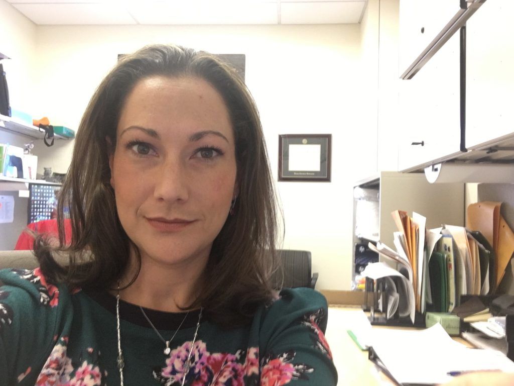 A woman in a floral shirt is taking a selfie in an office.