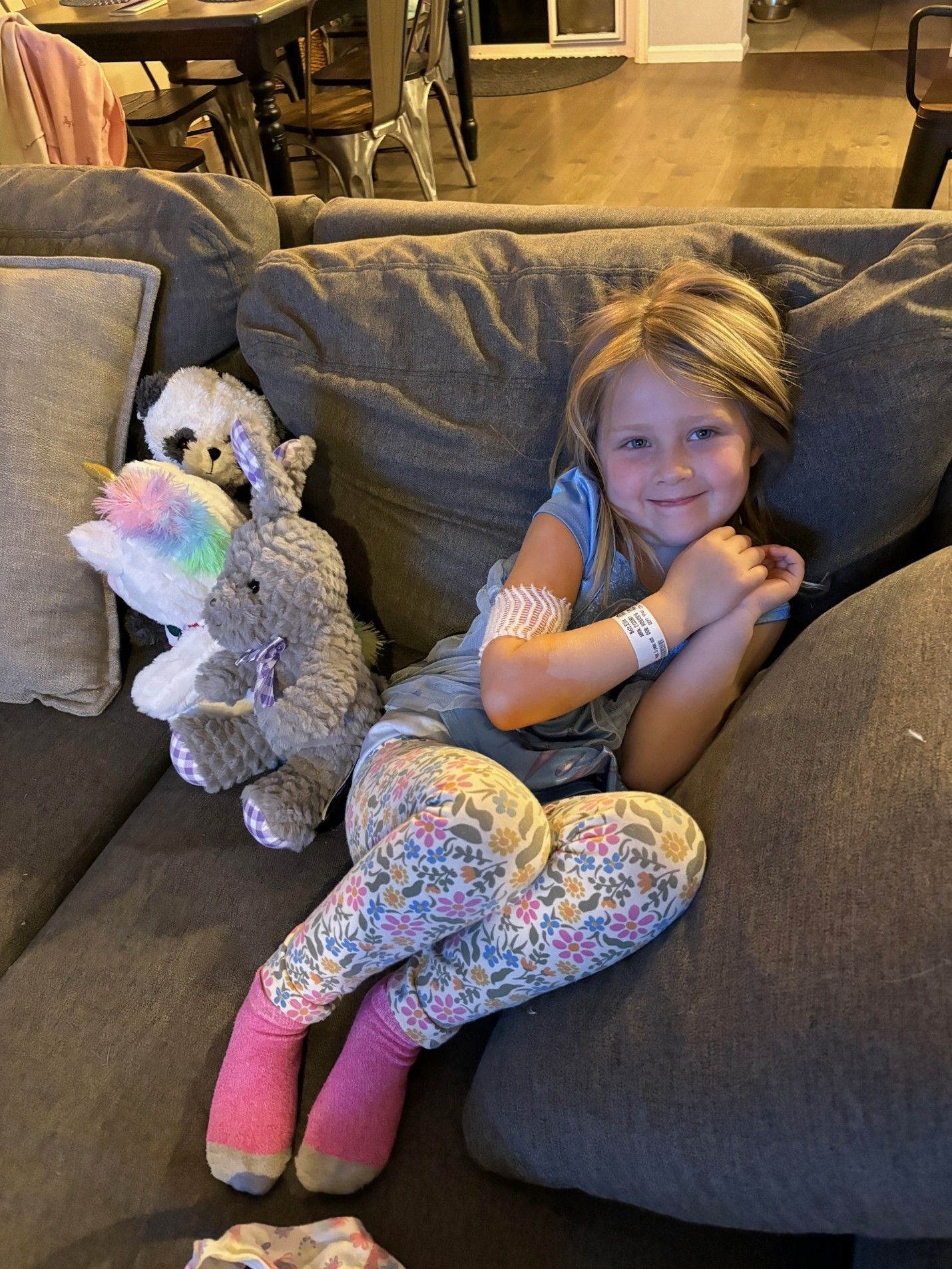 Five year old Ellie on the couch smiling with a bandage and hospital bracelet on her arm and wrist