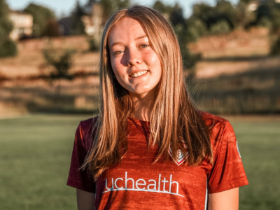 A young woman wearing a red shirt that says uchealth is standing in a field.