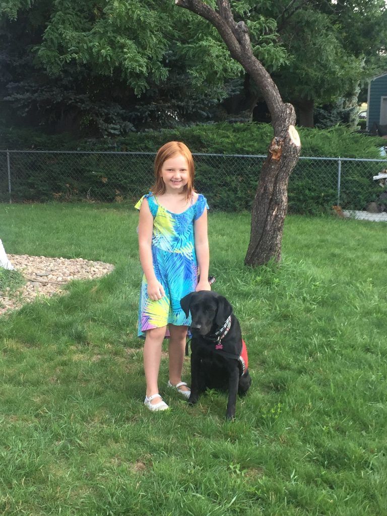 A little girl is standing next to a black dog in the grass.
