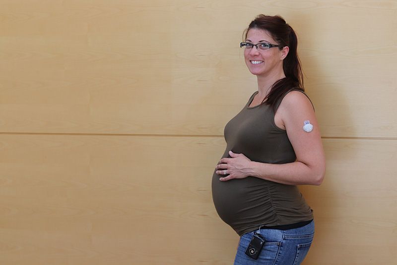 A pregnant woman with a patch on her arm is standing in front of a wall.