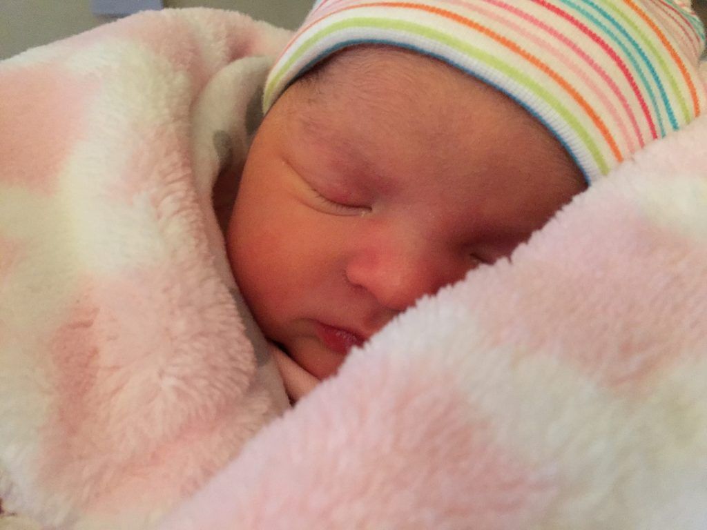 A newborn baby wearing a hat is sleeping on a pink blanket.