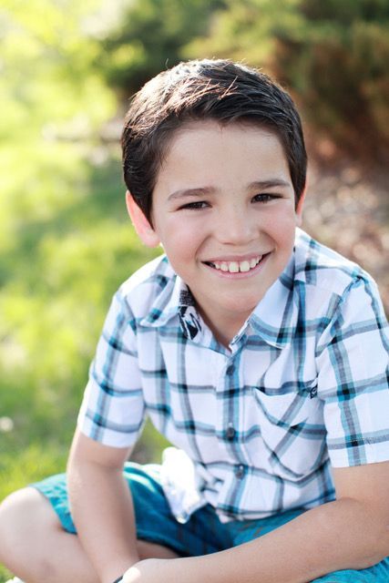 A young boy wearing a plaid shirt and blue shorts is sitting in the grass and smiling.