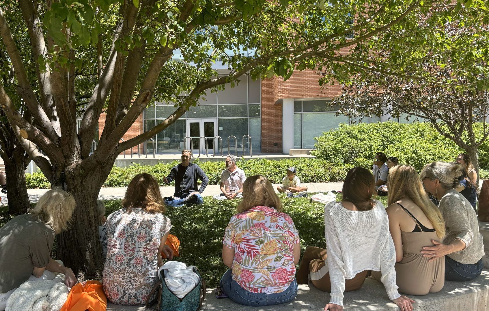 Group of people sitting outside under a tree listening to counselors.