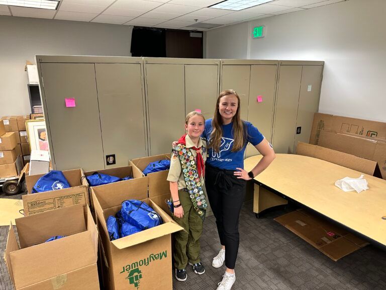 A girl scout and a woman are standing next to each other in a room filled with boxes.