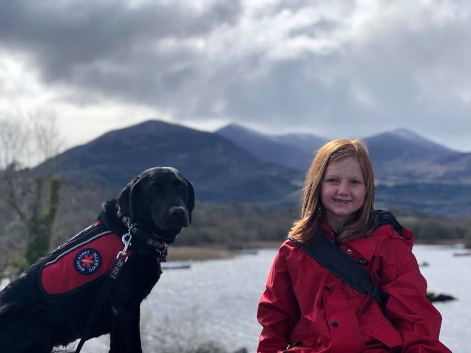 A girl in a red jacket is sitting next to a black dog.