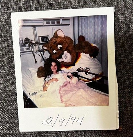 Author Mary Ann in hospital bed in 1994 smiling with team mascot