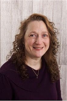 A woman with curly hair is smiling and wearing a purple sweater.