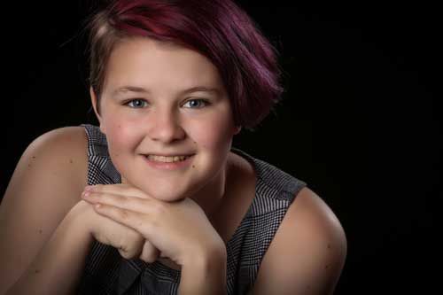 A young woman with purple hair is smiling with her hands on her chin.