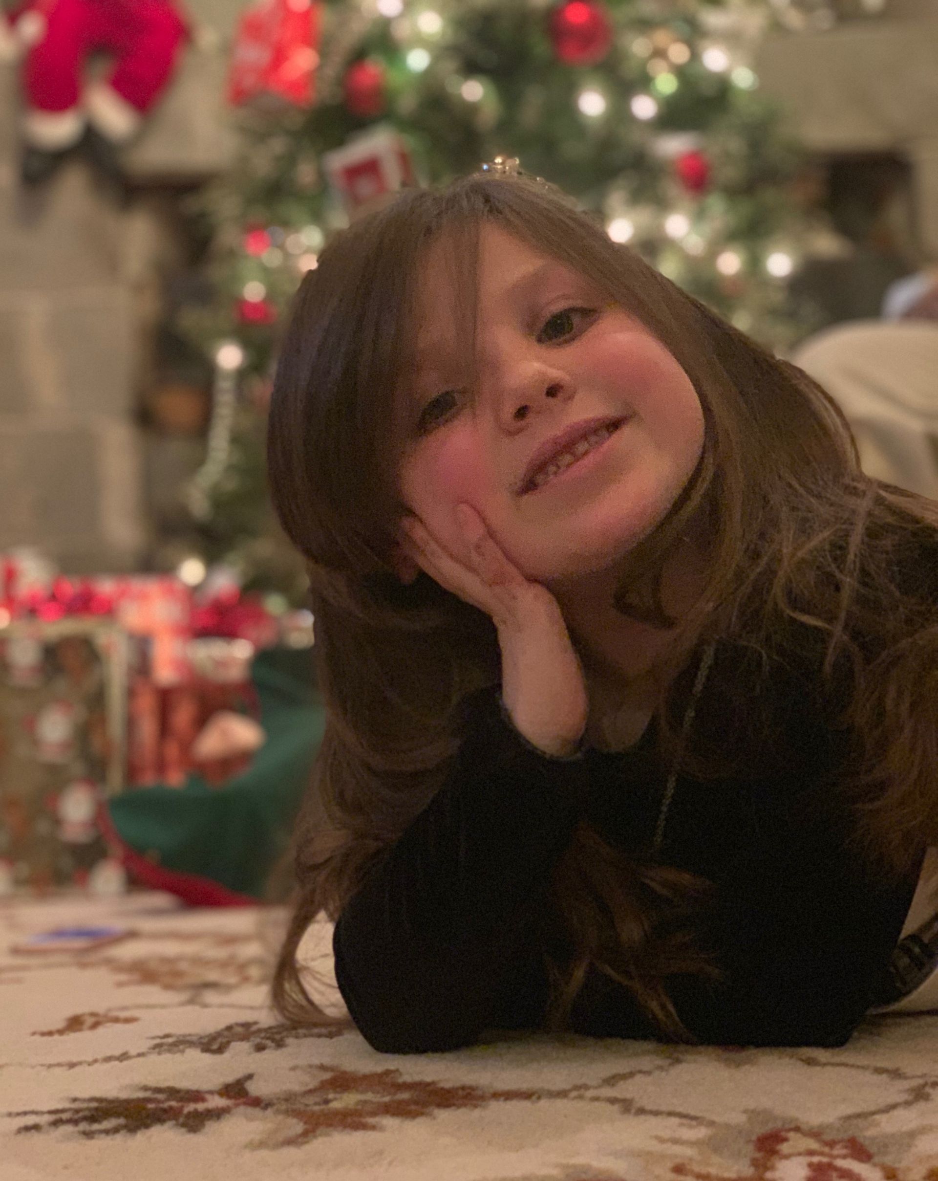 A little girl is laying on the floor in front of a christmas tree.