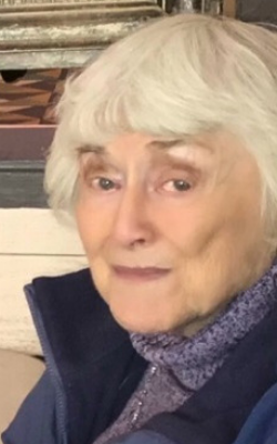 An elderly woman with white hair is wearing a blue jacket and a purple scarf.