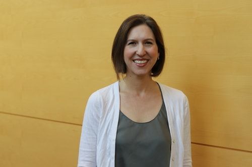 A woman in a white cardigan is smiling in front of a yellow wall.