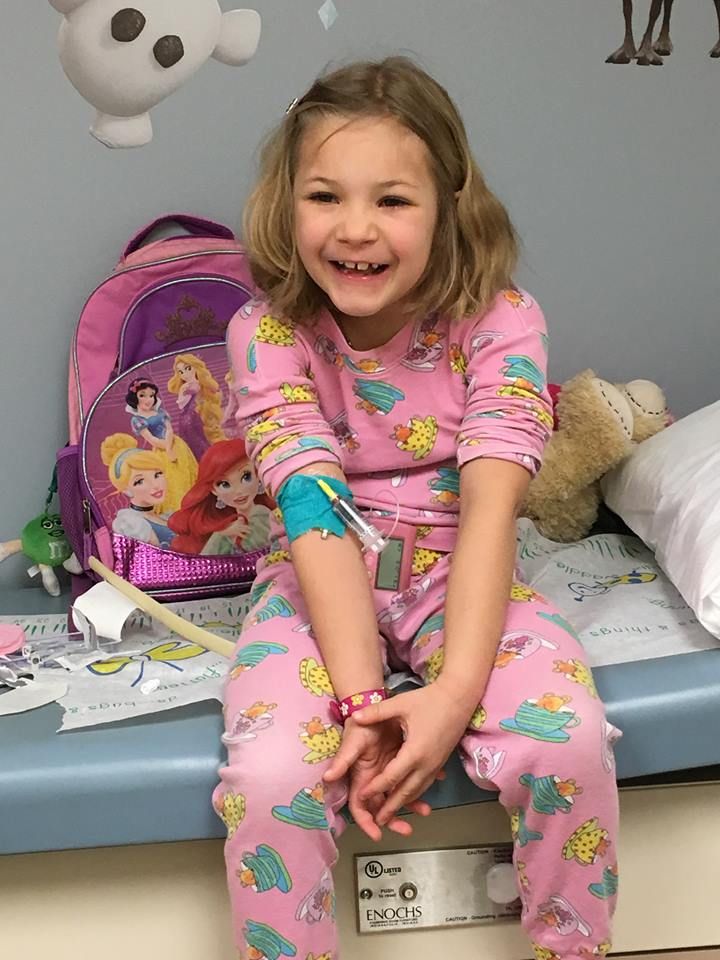 A little girl in pink pajamas is sitting on a couch with an iv in her arm.