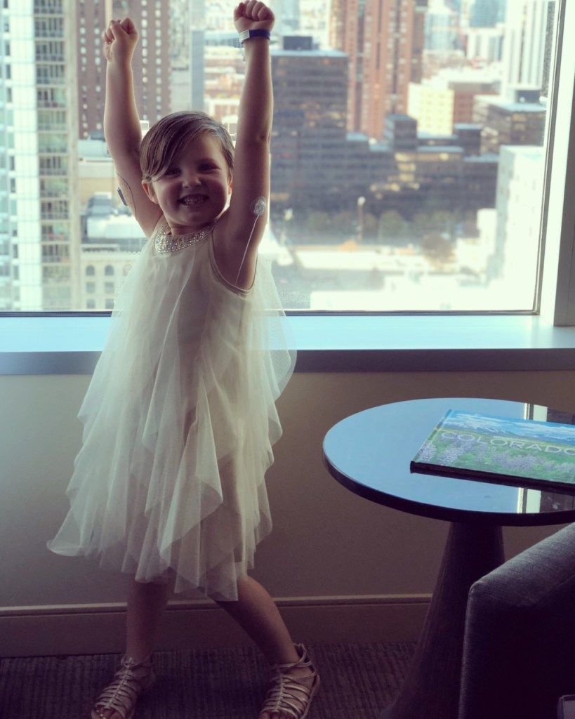 A little girl in a white dress is standing in front of a window with her arms in the air