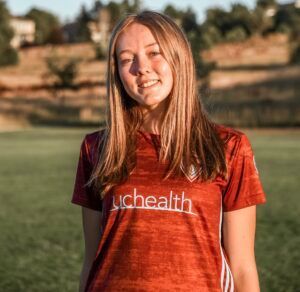 A young woman wearing a red shirt that says uchealth is standing in a field.