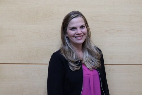 A woman in a pink shirt and black jacket is smiling in front of a wooden wall.