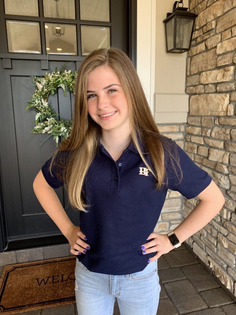A young girl wearing a blue polo shirt is standing in front of a door with her hands on her hips.