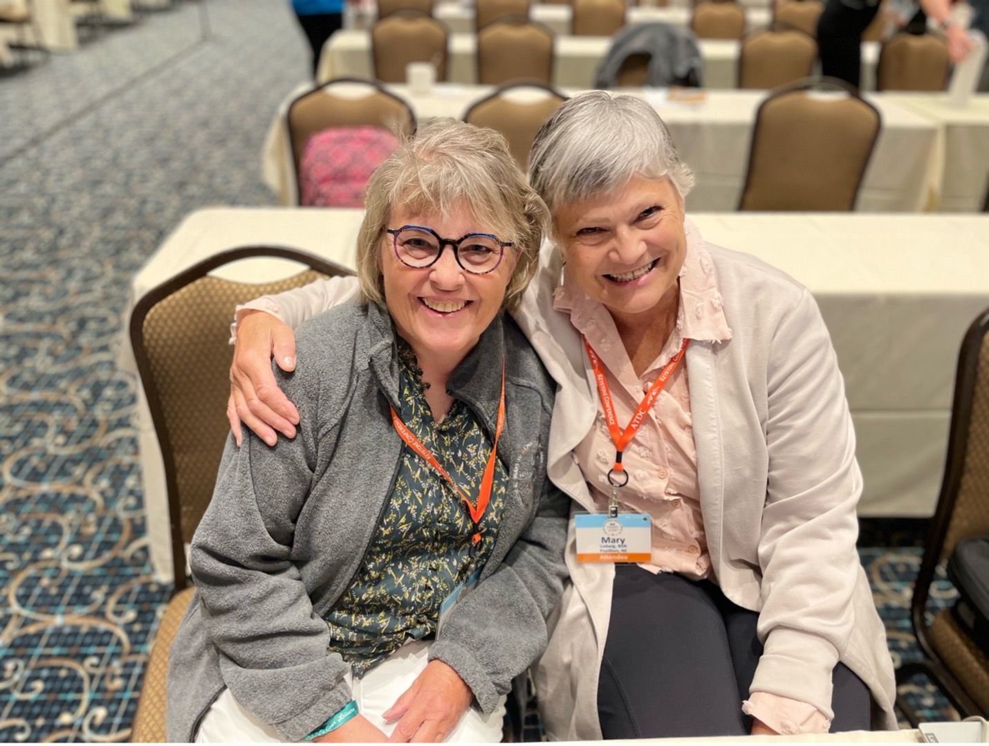 Two older female ATDC attendees sitting and smiling together with lanyards around their necks.